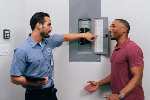 A Mr. Electric service professional holding a tablet pointing at an electrical panel and talking to another man.