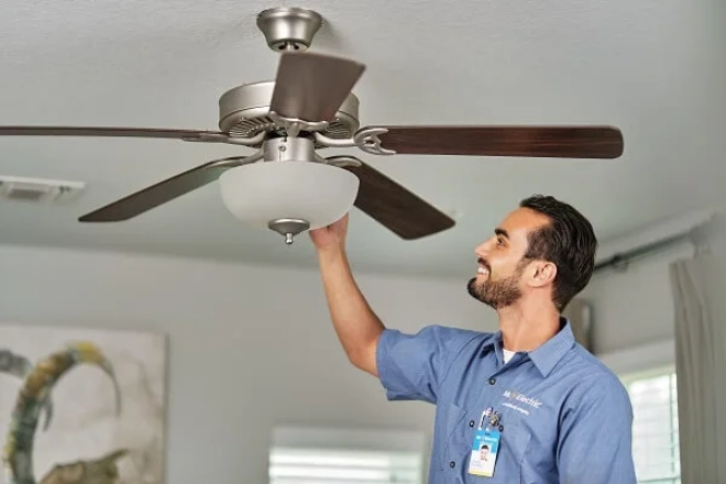 A smiling Mr. Electric electrician reaches over a light fixture attached to a ceiling fan